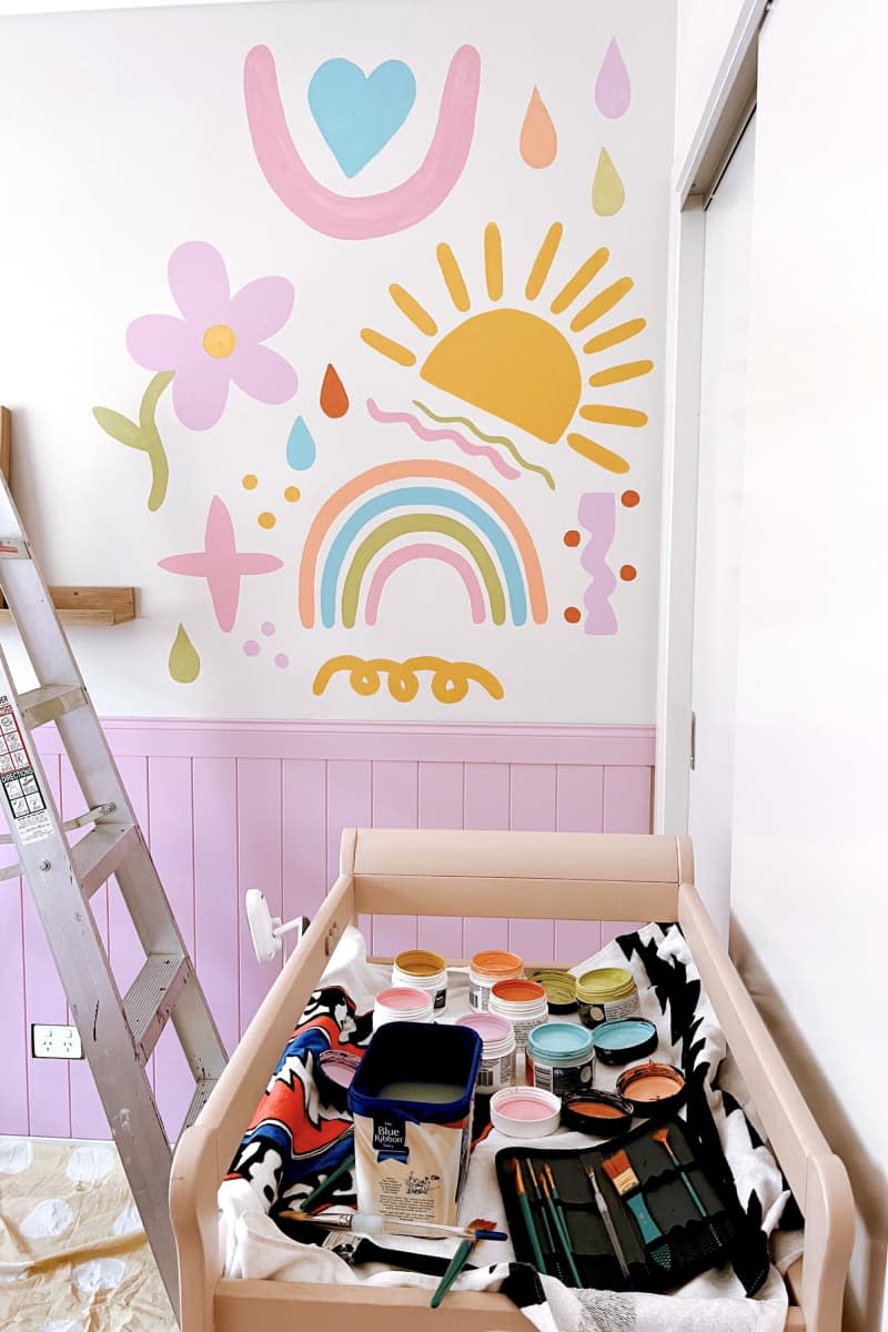 Wood cart with various paints, pink tongue and groove paneling up half wall, ladder, rainbow, sunshine, and flower doodles painted on wall
