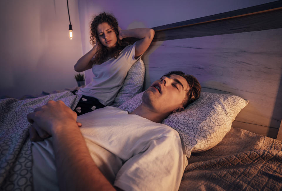 A woman sits in bed covering her ears as a man lies next to her, asleep and snoring