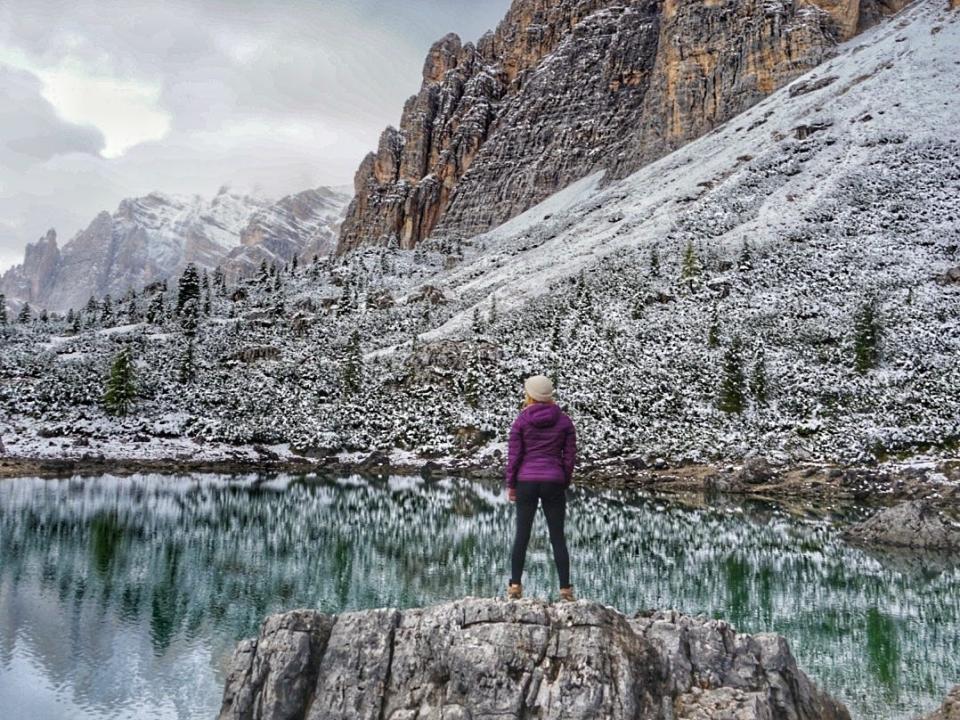 A woman standing on a rock in the middle of a lake surrounded by mountain and snow.