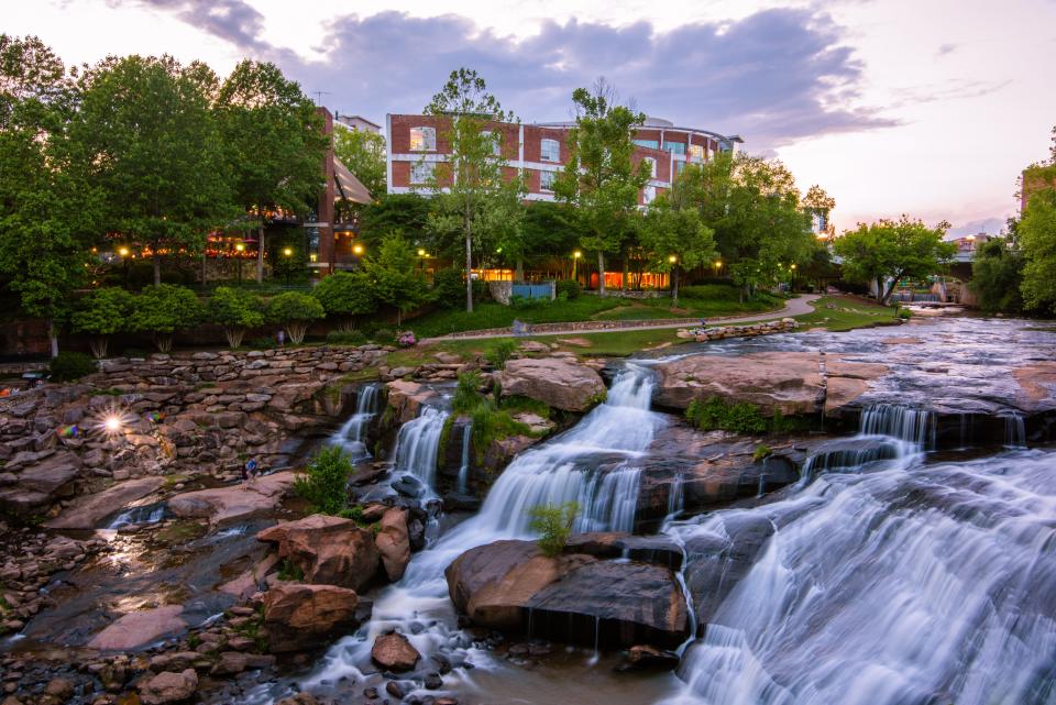 A waterfall at dusk with various rocks in front of a walking path, trees, and buildings. 