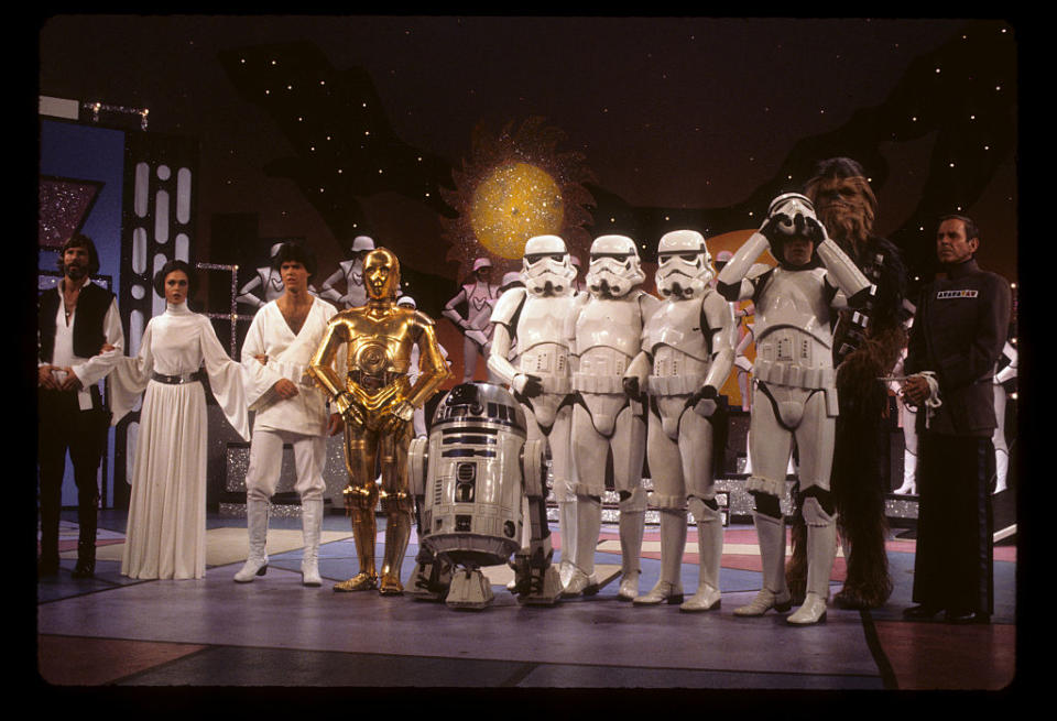 The cast of Star Wars, including Luke Skywalker, Princess Leia, C-3PO, R2-D2, Stormtroopers, Chewbacca, and others, posing together on a set