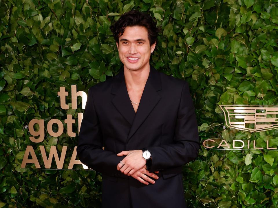 charles melton standing in front of a leaf background in a suit, smiling