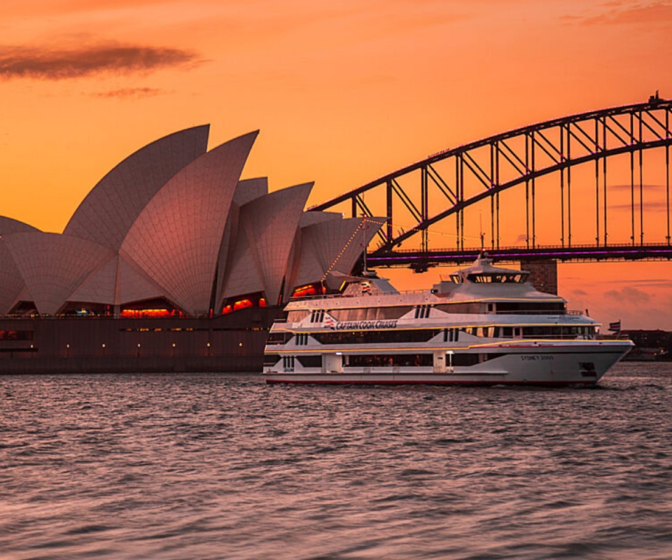 A white cruise ship on the Sydney Harbour at sunset with an orange sky behind the Opera House