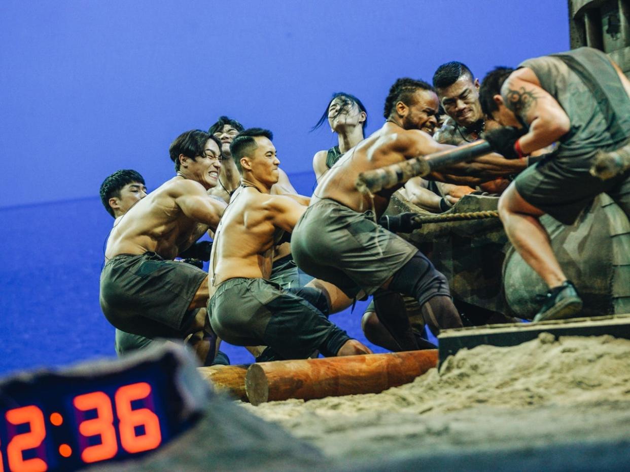 contestants on physical 100, many of them men not wearing shirts, strain to pull a large wooden boat across a series of logs on the sand