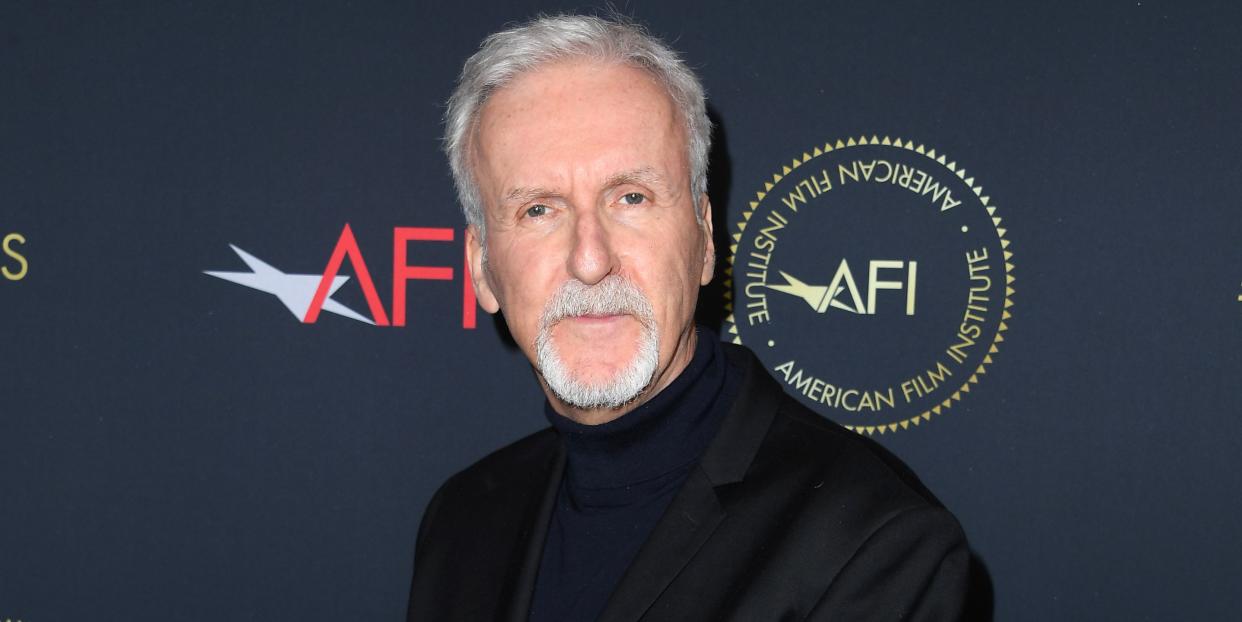 james cameron, an older man stands looking at the camera clasping his hands, he wears a black suit and turtleneck