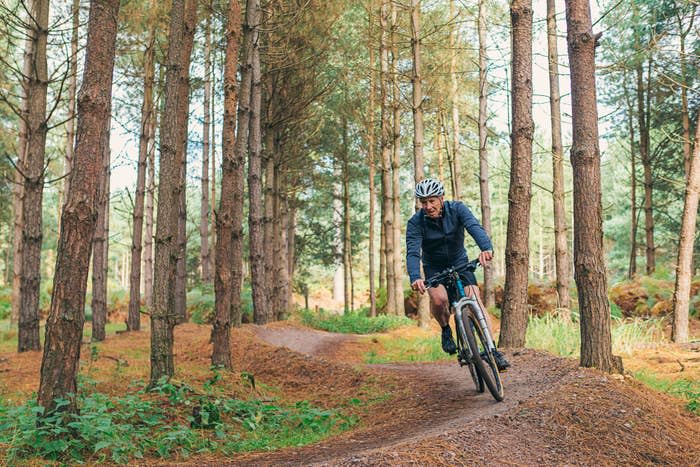 A person riding a mountain bike on a dirt trail through a forest, wearing a helmet and athletic clothing. The trail is surrounded by tall trees