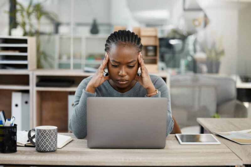 A woman sits, looking at her laptop and rubbing her temples as if stressed.