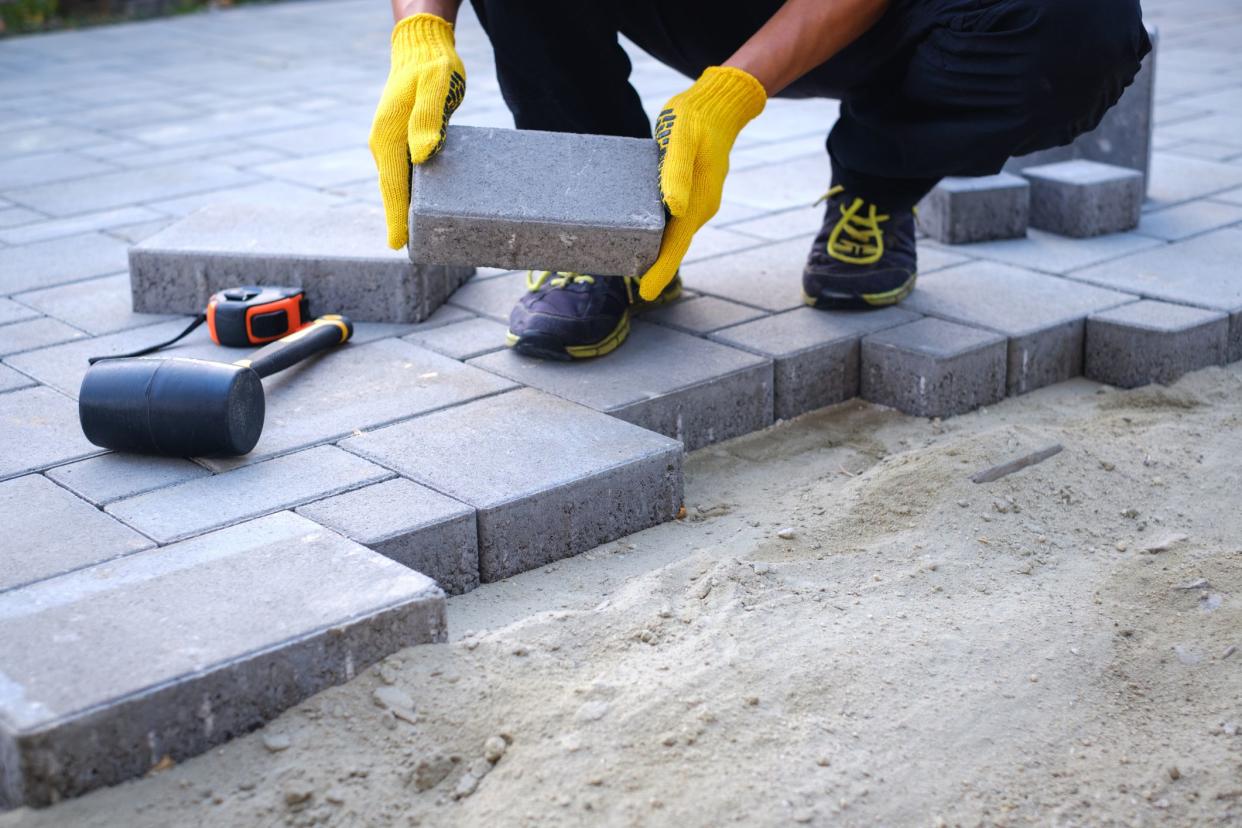 The master in yellow gloves lays paving stones in layers. Garden brick pathway paving by professional paver worker. Laying gray concrete paving slabs in house courtyard on sand foundation base.