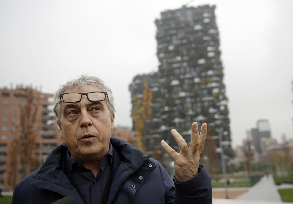 In this picture taken on Thursday, Nov. 15, 2018 architect Stefano Boeri gestures during an interview with the Associated Press as the vertical forest residential towers are visible in background, in Milan, Italy. If Italy's fashion capital has a predominant color, it is gray not only because of the blocks of uninterrupted neoclassical stone buildings for which the city is celebrated, but also due to the often-gray sky that traps in pollution. The city has ambitious plans to plant 3 million new trees by 2030_ a move that experts say could offer relief to the city’s muggy and sometimes tropical weather. (AP Photo/Luca Bruno)