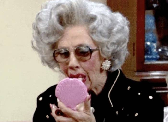 An elderly lady with curly hair, wearing sunglasses and pearl earrings, humorously applies lipstick while looking into a pink handheld mirror