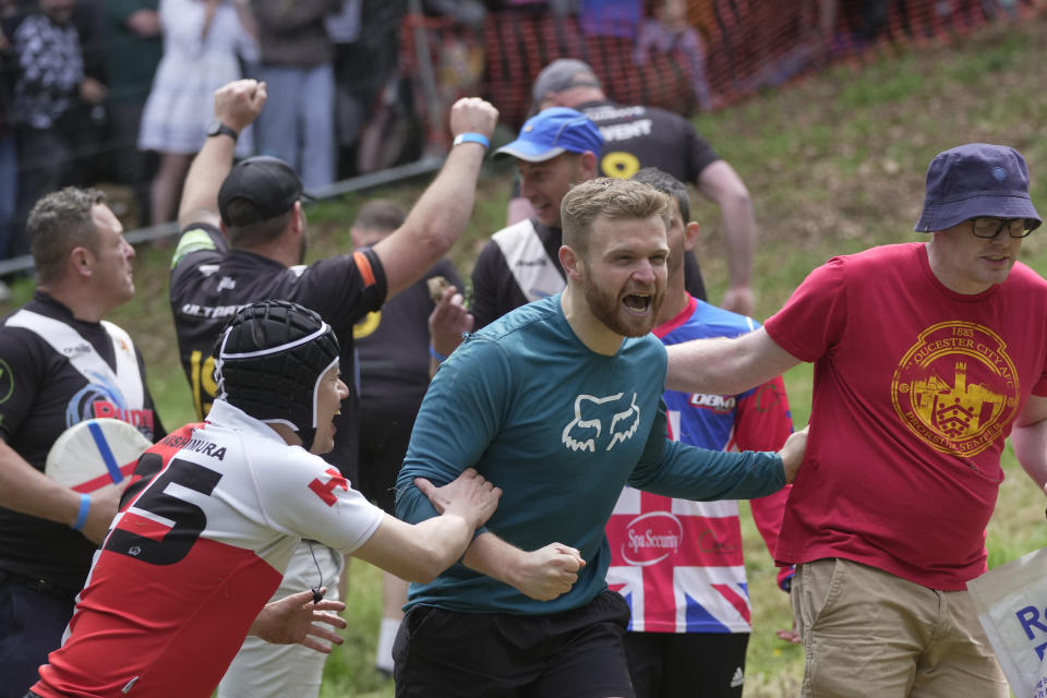 Matt Crolla, center, celebrates after winning the men's downhill race during the Cheese-Rolling contest at Cooper's Hill in Brockworth, Gloucestershire, Monday, May 29, 2023. The Cooper's Hill Cheese-Rolling and Wake is an annual event where participants race down the 200-yard (180 m) long hill chasing a wheel of double gloucester cheese. (AP Photo/Kin Cheung)