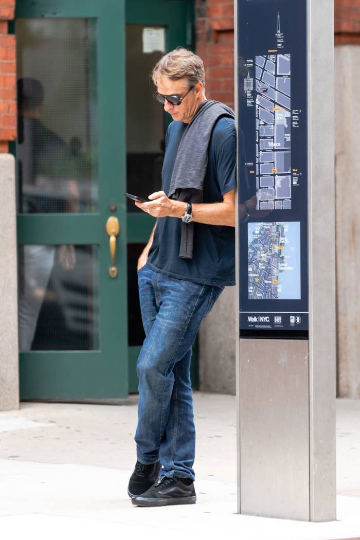 Tony looking at his phone as he leans up against NYC map sign post on a sidewalk