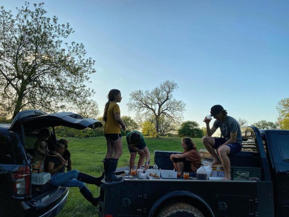 Joanna and her five children silhouetted having food stood on the back of a truck and sat in the trunk of the car