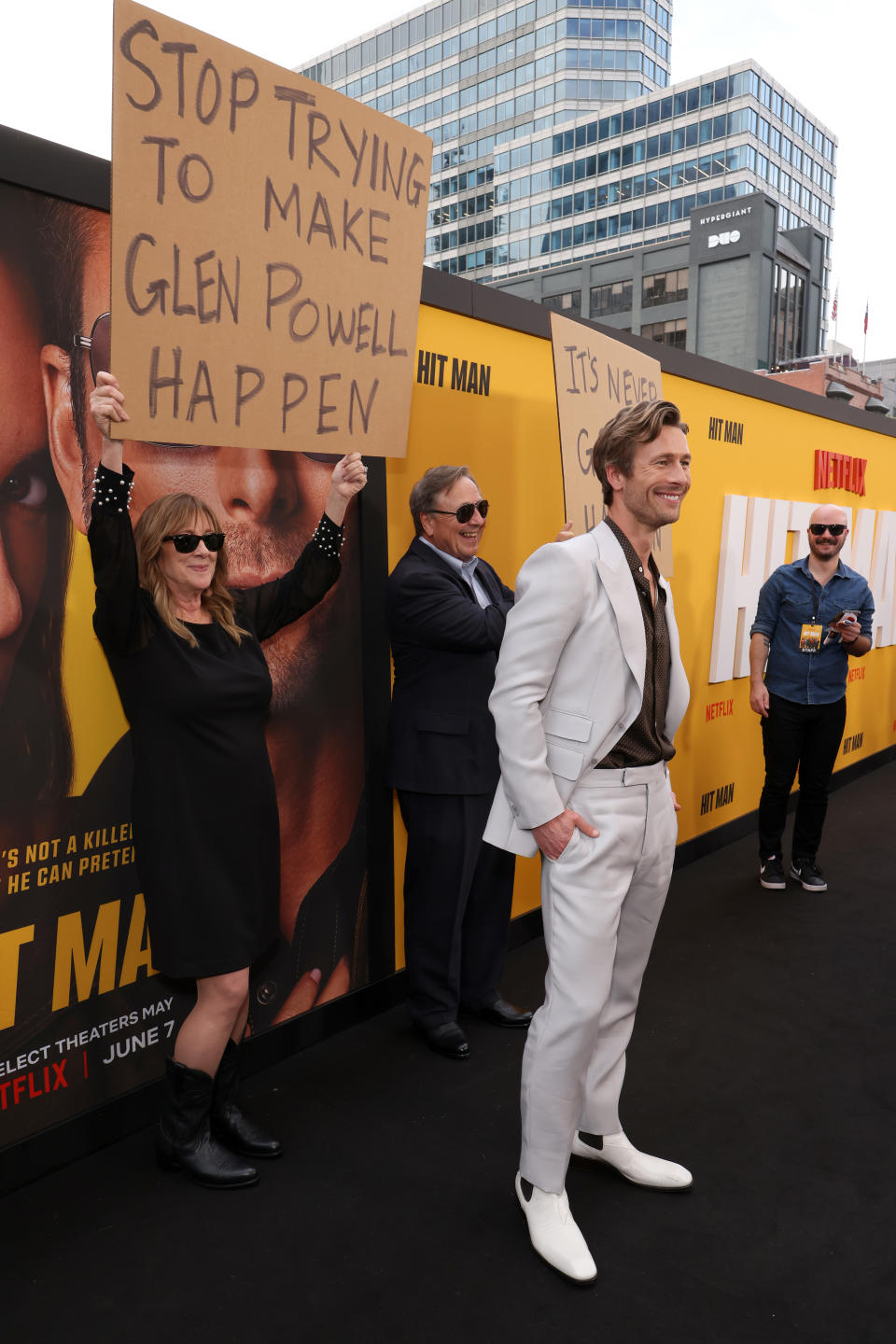 Glen Powell smirking in a light suit, standing before a poster with people holding signs, one reading "Stop trying to make Glen Powell happen."