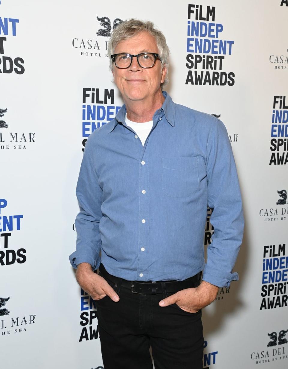 todd haynes smiles at the camera while standing in front of a white photo background with logos, he wears glasses, a blue collared shirt over a white tshirt and black pants