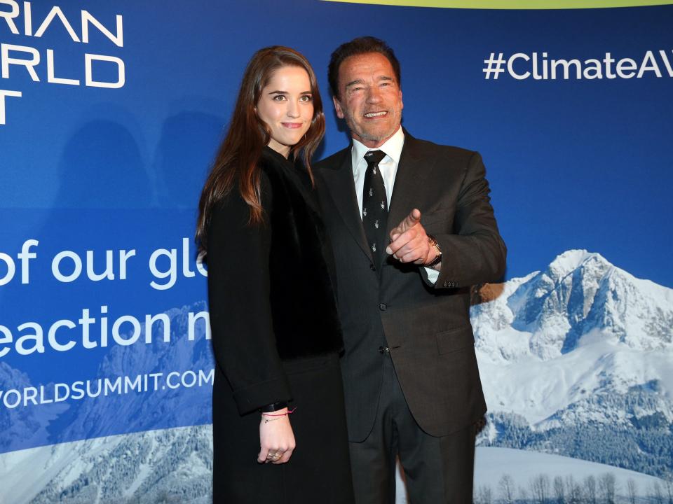 Arnold Schwarzenegger, who is pointing with his finger, and his daughter Christina Maria Aurelia Schwarzenegger standing together in front of a poster of mountains