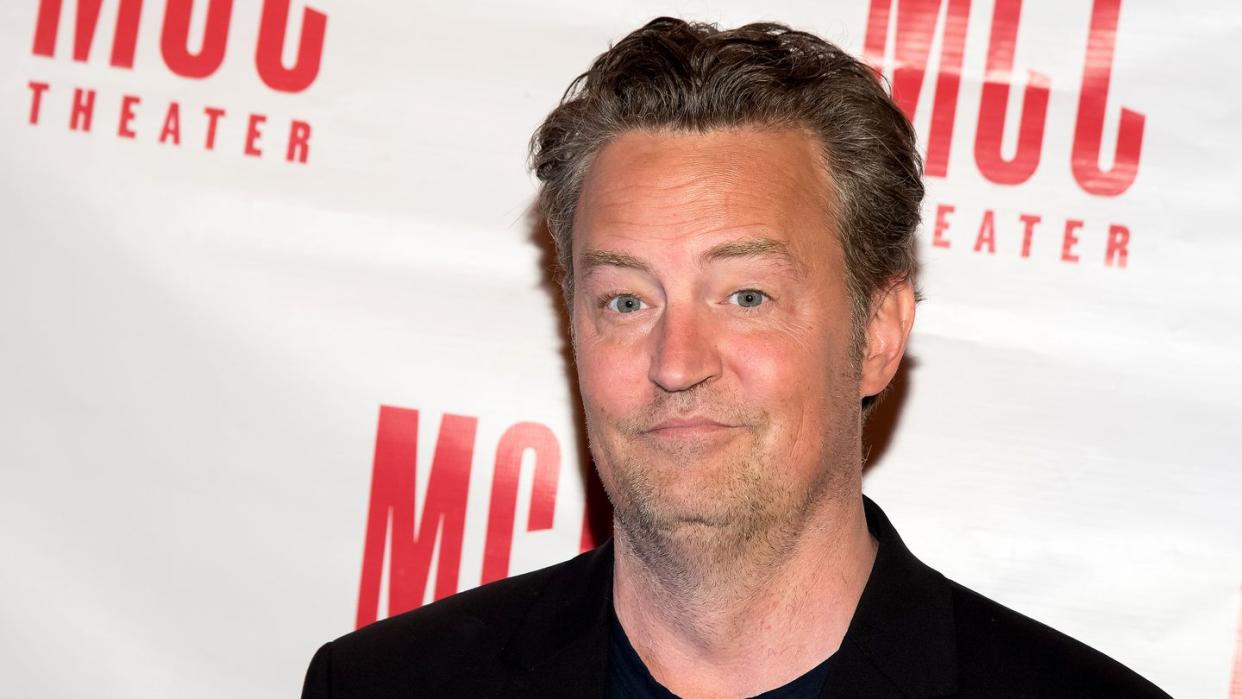 matthew perry shyly smiles while standing in front of a white background with red logos, he wears a black jacket over a black shirt