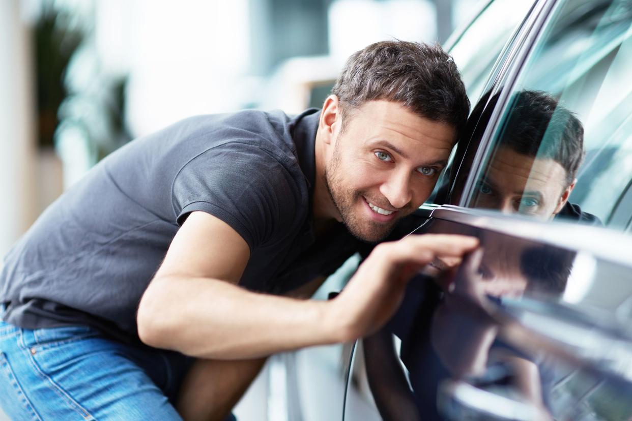Smiling man closely inspecting exterior driver's side of a dark colored car in a dealership showroom, blurred background