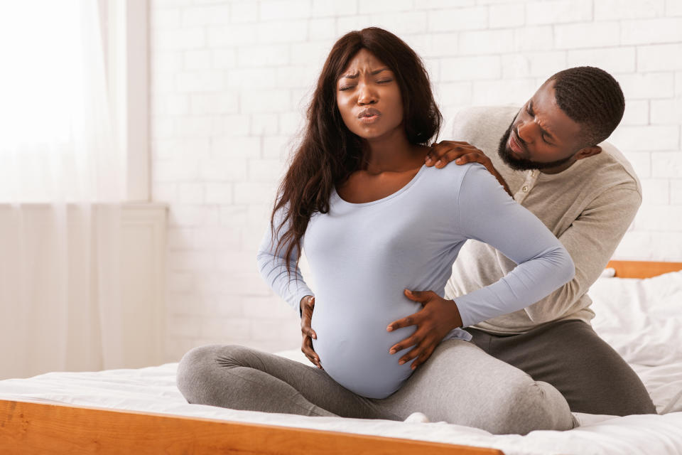 Pregnant black lady suffering from back pain, sitting on bed, her attentive husband massaging her shoulders, free space