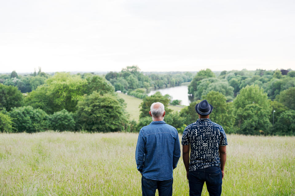 A man in a denim shirt and another in a patterned shirt and hat stand side by side, looking at a scenic view of a river and trees