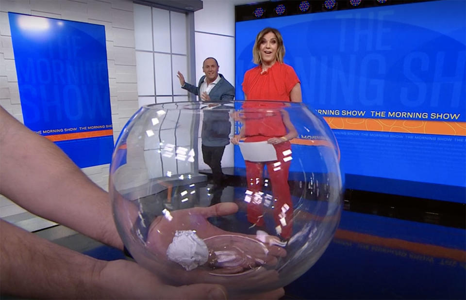 Larry Emdur and Kylie Gillies looking surprised with a fishbowl in front of the camera holding a paper ball