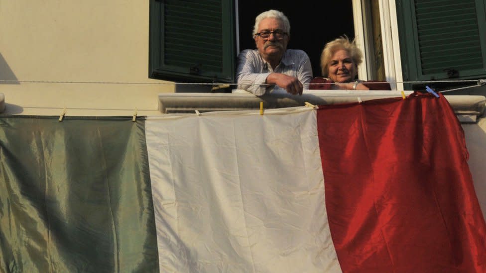 Una pareja mayor en un balcón con una bandera italiana.