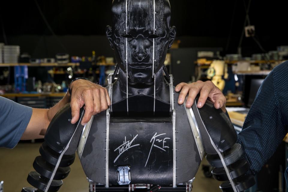 Two human hands resting on the shoulders of a humanoid robot marked with signatures