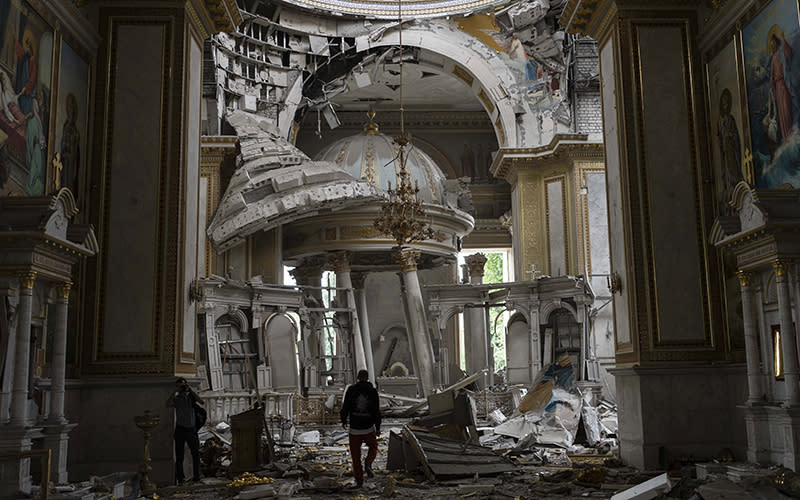 Church personnel inspect damages inside the Odesa Transfiguration Cathedral. One person is shown very small in a large room. There is a great deal of rubble on the ground and structures and columns on the altar are skewed and tilted. Parts of the decoration on the upper walls and ceiling are peeling and hanging down.