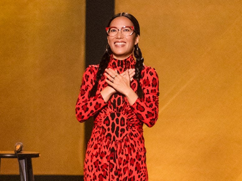 A women with long dark hair in pigtails and a red animal print dress stands with her arms crossed over her chest, smiling as a the crowd cheers.