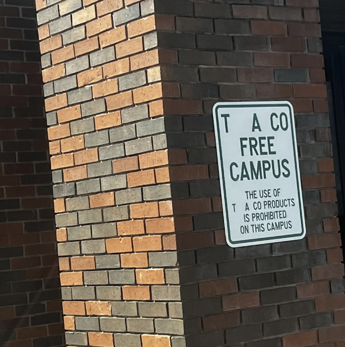 Sign on brick wall stating campus is tobacco-free, banning use of such products, but someone made it spell "TACO"