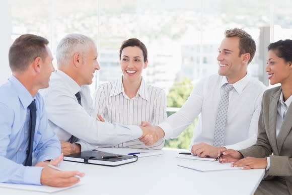 A group of professionally-dressed people sitting around a table smiling and shaking hands.