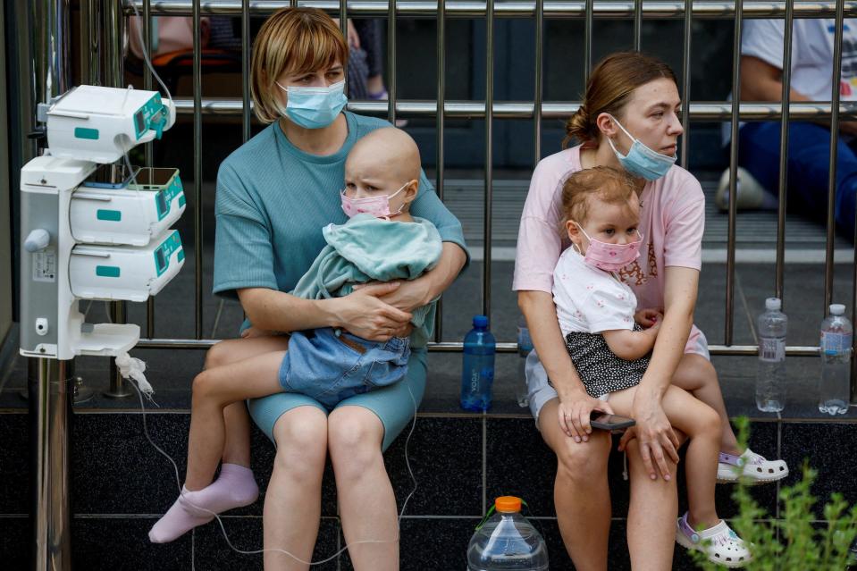 Two women sit and hold young children on their lap. One child has tubes in their body