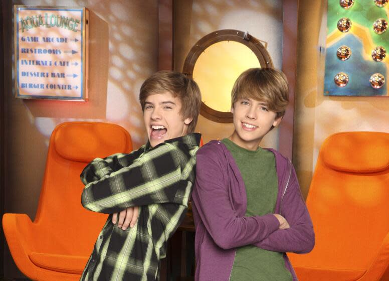 Dylan Sprouse in a flannel shirt with his arms crossed leaning against Cole Sprouse in a purple jacket and a green shirt.
