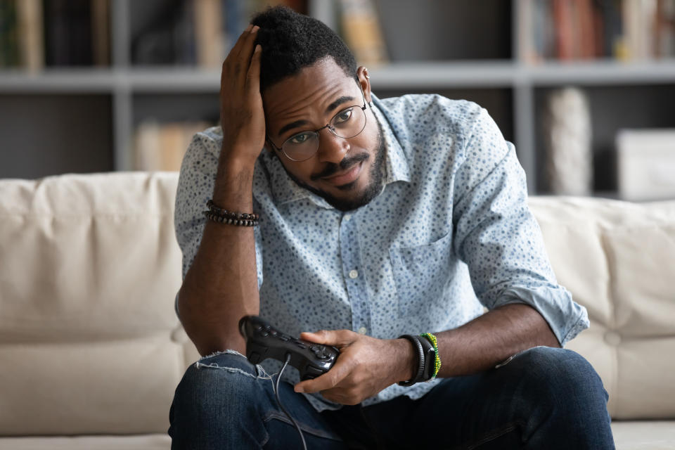 Unhappy African American gamer player dissatisfied with video game losing, upset young man wearing glasses holding gamepad controller joystick, sitting on couch in living room, using console