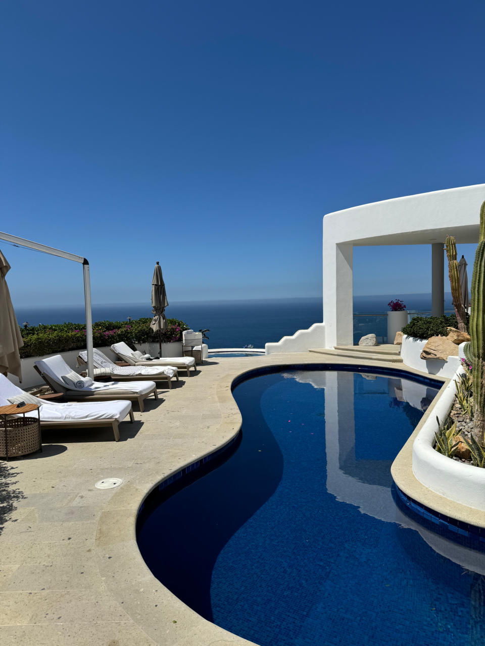 A luxurious, curved poolside area with lounge chairs and umbrellas overlooks a serene ocean view, adjacent to white modern architecture and cacti