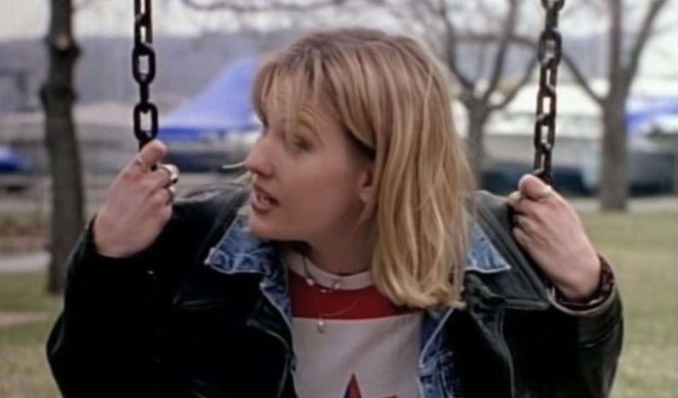 Joey Lauren Adams sits on a swing outside. She looks sideways and is upset.