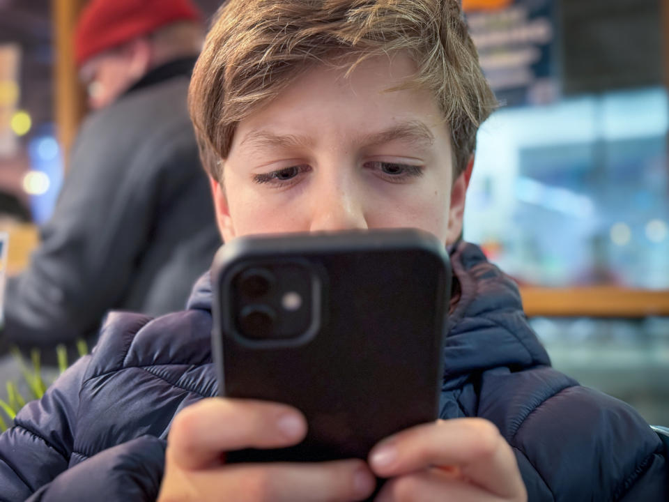 A young boy concentrates on a smartphone while sitting indoors, wearing a puffy jacket. An adult in a red hat is visible in the background