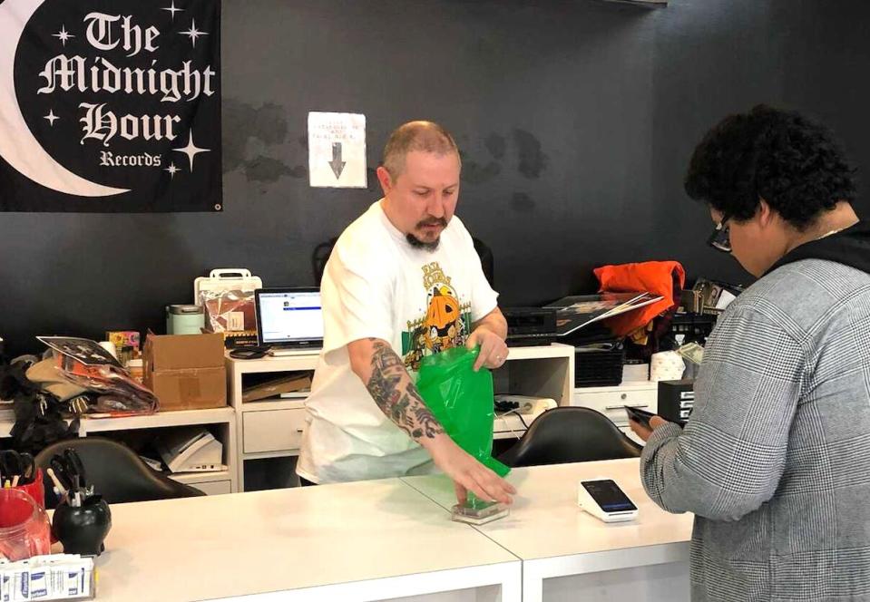 A man helps a customer make a purchase at a store counter, with a logo reading "The Midnight Hour Records" behind him