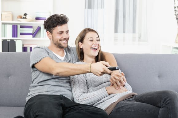 Couple cuddling on the couch smiling watching TV with man holding the remote.