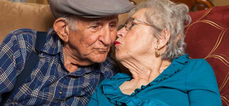 An elderly woman kisses her elderly husband on the cheek.