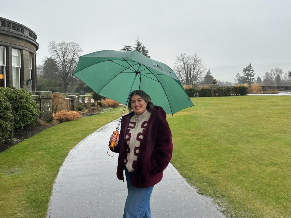 A girl smiling while holding an umbrella in front of green fields.