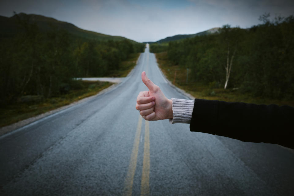 Person hitchhiking by raising thumb on a rural road with trees and hills in the background