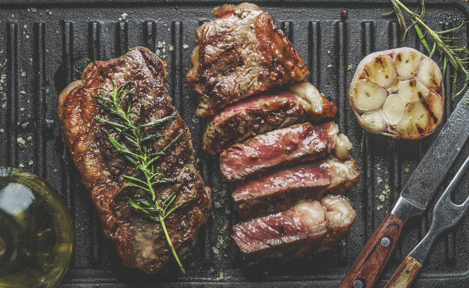 Close up of sliced roasted medium rare barbecue steak with rosemary, roasted garlic and cutlery on rustic iron grill. Cooking at home with fresh meat and flavorful herbs. Top view.