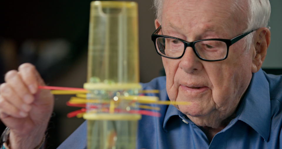 Goldfarb demonstrates one of his most successful games. KerPlunk is a childrens game involving a plastic tube filled with small marbles that rest on top of interlaced sticks. Players take turns removing them one by one, trying to avoid making any marbles fall.