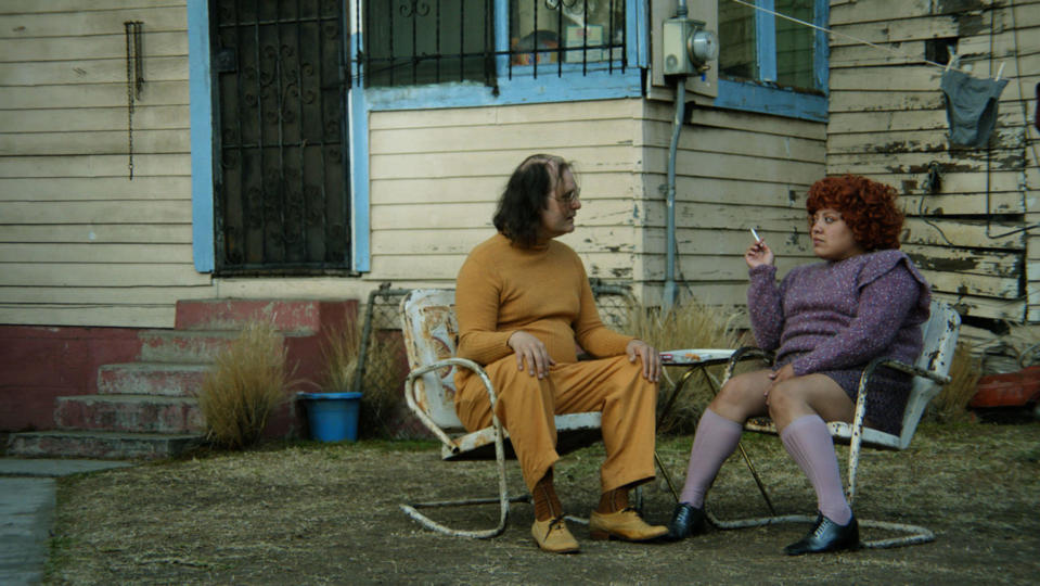 A bespectacled man in a yellow tracksuit sits next to a woman in a purple dress and large curly hair on a dilapidated lawn