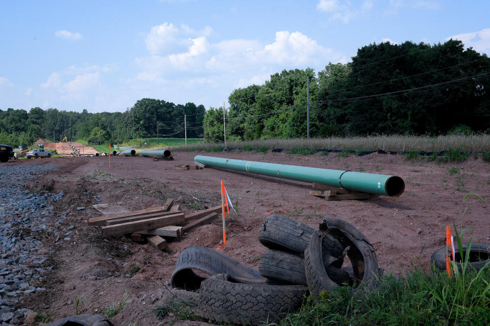 A long line of green pipe segments stretches across a ribbon of recently excavated dirt, with a few orange markers and a heap of used tires in the foreground.