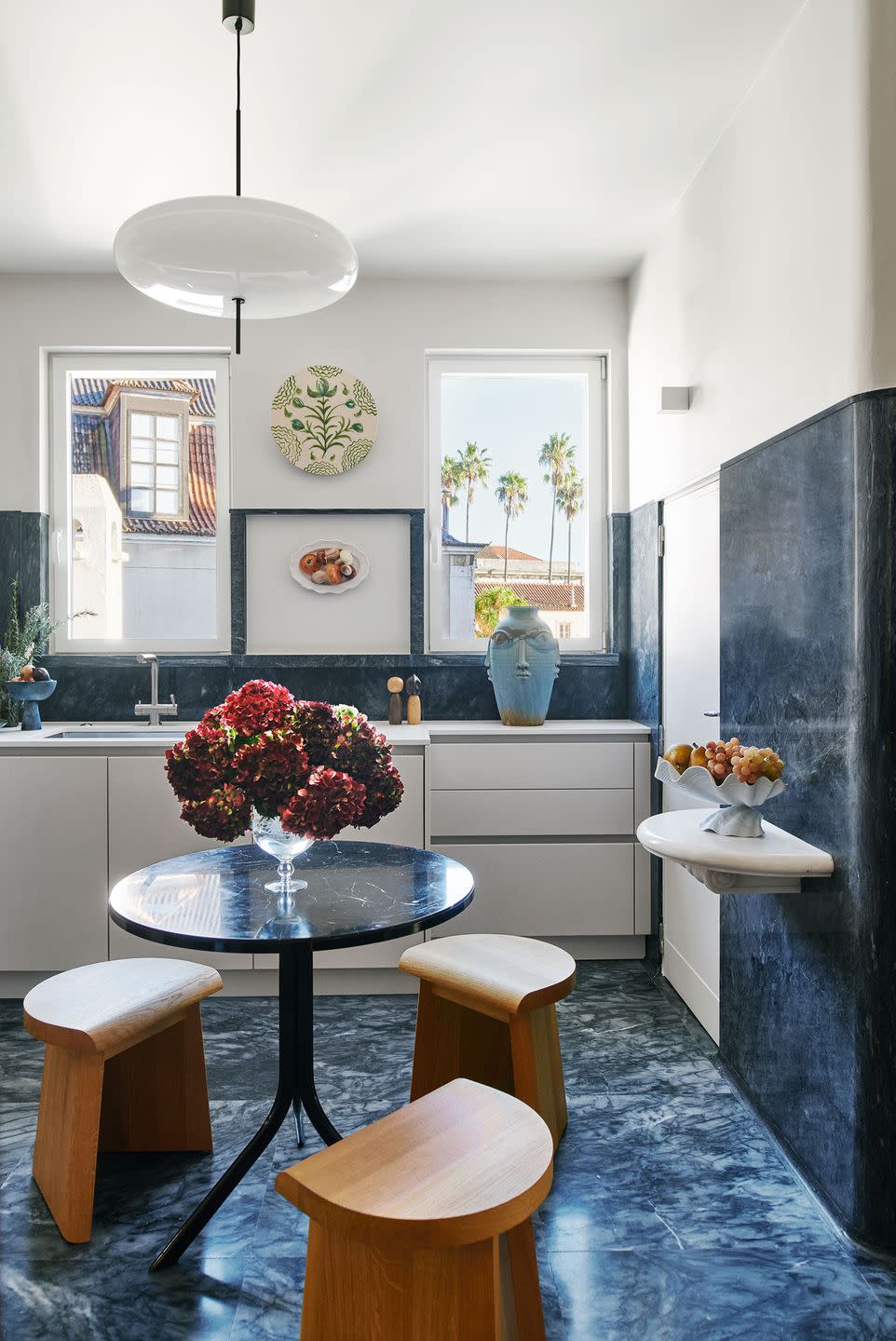 a kitchen’s floor, backsplash, partial wall, and a round table are a blue marble, three stools are oak, two windows are above white cabinets and sink, a floating shelf has a fruit bowl, a spheroid pendant above table