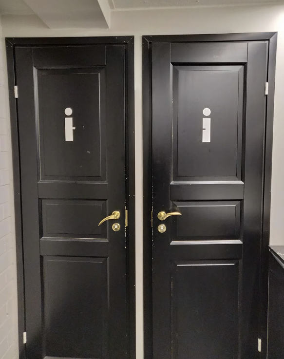 Two black restroom doors, each with a simple sign depicting a standing figure. The figure on each door is not gender-specific