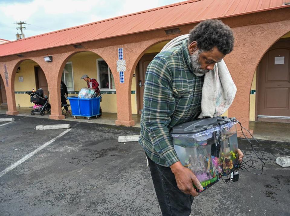 Michael Williams, 52, carries a fish tank on Feb. 15 that had been left outside overnight by property managers at The Greens Hotel with their beloved pet betta fish Bluez. The family of four had been locked out of the room the night before. All their belongings were still locked inside the room including his medication for chronic pulmonary disease. In the background his wife searches a garbage bin for their belongings.