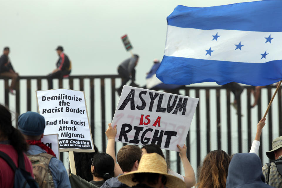 Pro-migrant caravan demonstrators, including a woman waving a Honduran flag, rally&nbsp;as some people climb the border wall.